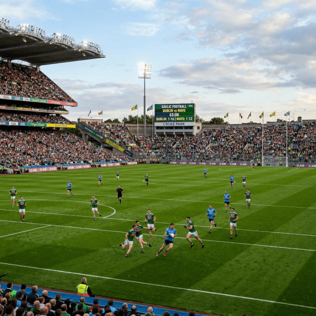 Gaelic football players from Dublin and Mayo competing on a green pitch with a large crowd in stands